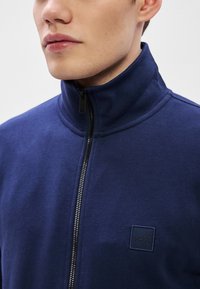 Tumši zils rāvējslēdzēja džemperis no mīksta auduma ar augstu apkakli un kvadrātveida BOSS logotipu krūtīs, ar gludu tekstūru.