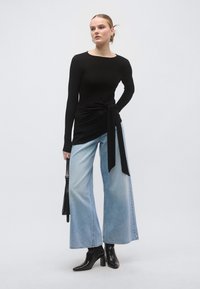 Top negru cu mâneci lungi și detaliu cu nod, asortat cu blugi evazați bleu deschis și ghete negre de gleznă, creând o siluetă modernă.