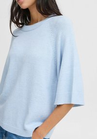 Světle modrý svetr s krátkým rukávem, kulatým výstřihem a strukturovaným prošíváním podél ramenních linií. Volný střih. Měkká pletená látka.