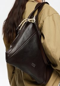 Ciemnobrązowa skórzana torba o trójkątnym kształcie, z plecionym akcentem w kolorze kremowym i czarnym w paski oraz wytłoczonym logo.