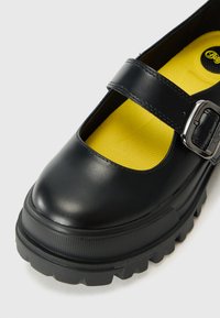 Scarpa Mary Jane in pelle nera con punta rotonda, cinturino con fibbia e suola in gomma nera testurizzata. L'interno presenta una soletta gialla brillante.