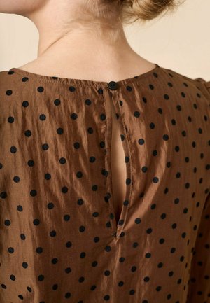 Brun stoftop med sorte polkaprikker; har en knaplukning bagpå og en split-udskæring. Sildestruktur med en let draperet udseende.