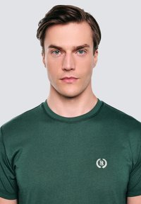 Tamsiai žalia trumpa rankovė t-shirt, pagaminta iš minkštos medžiagos, su mažu baltu logotipu ir lauro akcentais ant krūtinės bei apykakle. 