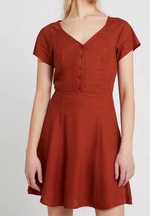 Femme portant une robe à manches courtes couleur rouille avec un décolleté en V et des boutons sur le devant, debout devant un fond clair uni.