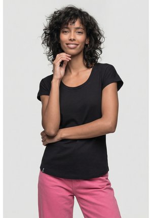 Frau mit lockigem Haar, die ein schwarzes Kurzarmshirt und pinke Hose trägt, lächelt, wobei eine Hand nahe am Kinn und die andere verschränkt ist.