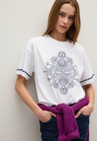 Witte katoenen t-shirt met een gedetailleerd marineblauw bloemenontwerp, korte mouwen met marineblauwe afwerking, gecombineerd met donkere jeans en een paarse gebreide trui om de taille.
