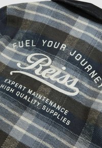 Mėlyna ir pilka langeliais marginta fliso medžiaga su balta siuvinėta tekstu: "Kurk savo kelionę" ir "Reiss", taip pat "Ekspertinė priežiūra, aukštos kokybės tiekimas".