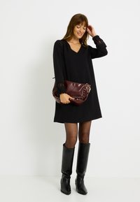 Robe noire à manches longues avec des accents en dentelle, assortie à une pochette bordeaux ornée de clous. Modèle portant des bottes noires jusqu'aux genoux.