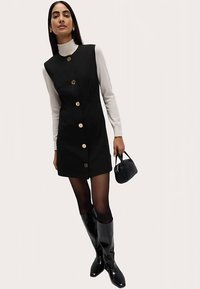 Zwarte mouwloze jurk met gouden knopen, gecombineerd met een wit shirt met lange mouwen, zwarte panty's en glanzende zwarte kniehoge laarzen. Kleine handtas.