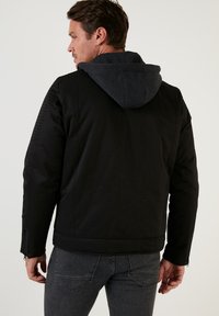 Svart jacka med huva, med texturerad tyg, figursydd design och dragkedjor vid ärmarna. Grå huva ger en kontrasterande detalj.