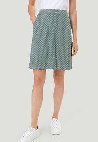 Femme portant une jupe longueur genou avec un motif géométrique, des baskets blanches et un haut blanc, se tenant avec une main dans la poche.