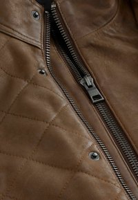 Veste en cuir marron avec une texture matelassée, fermeture éclair argentée proéminente et détails de boutons pression près du col.