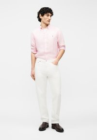 Άνδρας που στέκεται με το χέρι στην τσέπη, φοράει ροζ ριγέ μακρυμάνικο πουκάμισο, λευκό παντελόνι και σκούρα καφέ παπούτσια σε απλό φόντο.