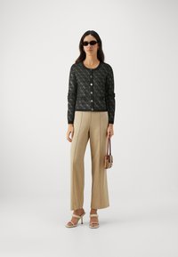 Black knitted cardigan with diamond pattern, round neckline, front buttons, paired with beige wide-leg trousers and white heeled sandals.