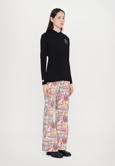 Top negro de manga larga con cremallera frontal y detalle de logo, combinado con pantalones de pierna ancha estampados que presentan imágenes y texto coloridos. Zapatos negros.