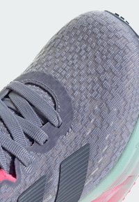 Gray athletic shoe with textured upper, flat laces, and purple accents. Features a patterned design and a contrasting mint outsole.