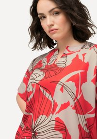 Bluse med blomsterprint i rød og beige med V-hals og korte ærmer, der har markante sorte linjer og en løs pasform.