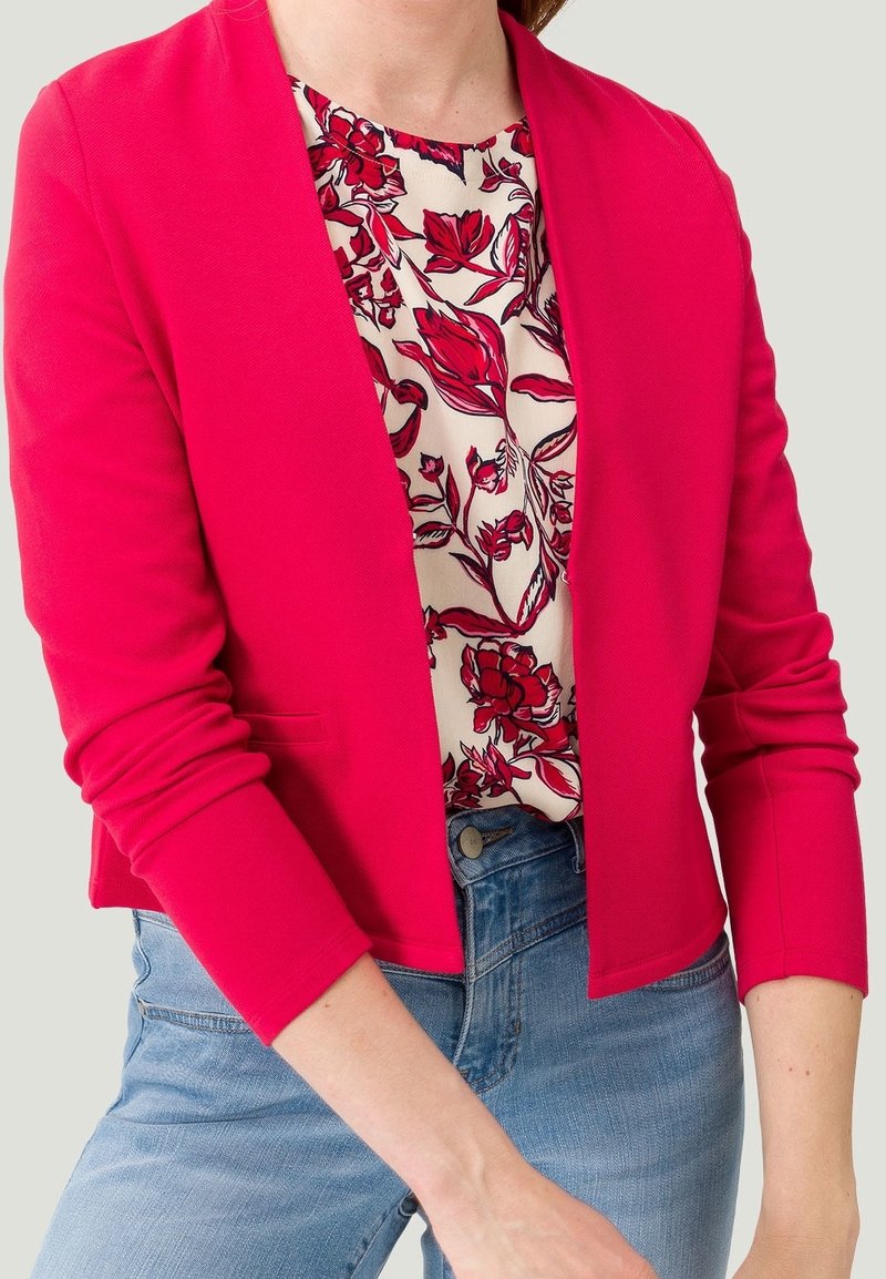 Femme portant un blazer rose vif sur un chemisier blanc à imprimé floral rouge et un jean bleu, cadrée à la taille avec les bras visibles.