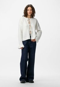 White knitted sweater with a cropped design and lace-up front, paired with dark blue flared pants and black shoes. Smooth texture.