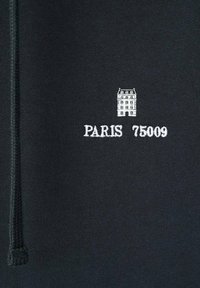 Schwarzer Pullover mit weißer Stickerei eines Gebäudes und dem Text "PARIS 75009." Das Material wirkt glatt mit einer strukturierten Oberfläche.