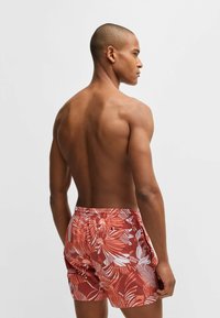 Men's swim shorts in rust red with white tropical leaf patterns, featuring an elastic waistband and a back pocket. Smooth, lightweight fabric.
