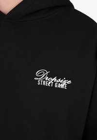 Hoodie noir avec le texte blanc "Dropsize Street Game" sur la poitrine, montrant la texture du tissu et les détails de couture de la capuche.