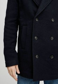 Marineblå ullfrakk med dobbeltspent design, store svarte knapper og sidelommer. Teksturen ser myk og strukturert ut. Synlig denim under.
