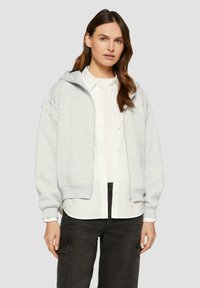 Hellgrauer Reißverschluss-Hoodie mit gerippten Bündchen, großer Kapuze und Verzierungen an den Ärmeln, getragen über einem weißen Hemd mit Knöpfen und dunklen Hosen.
