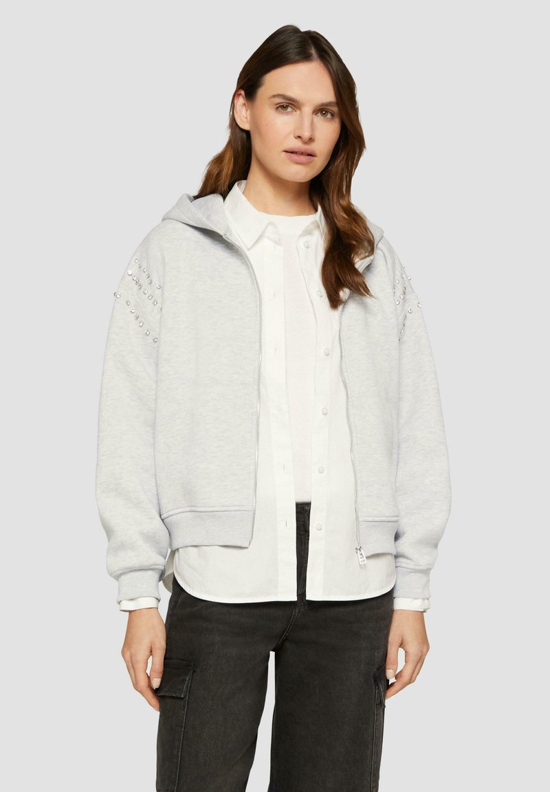 Hellgrauer Reißverschluss-Hoodie mit gerippten Bündchen, großer Kapuze und Verzierungen an den Ärmeln, getragen über einem weißen Hemd mit Knöpfen und dunklen Hosen.