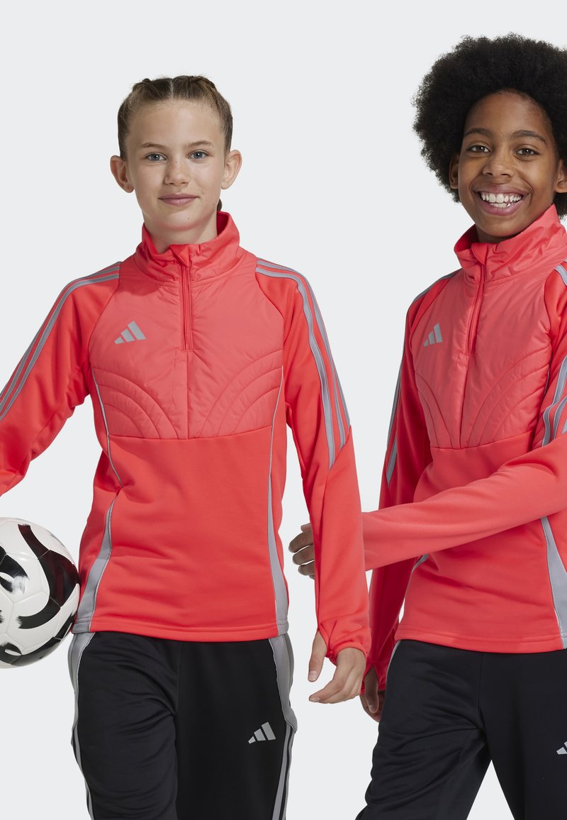Vestes de sport corail vif avec des rayures grises, des cols hauts et des détails matelassés à l'avant. Associées à un pantalon noir et un ballon de football.