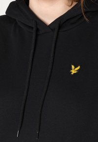 Un hoodie noir en tissu doux avec une poche kangourou. Il présente un logo brodé jaune et des cordons de serrage ajustables.