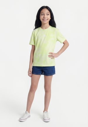 Dziewczyna z długimi ciemnymi włosami, ubrana w jasnozielony t-shirt, niebieskie dżinsowe szorty i białe trampki, stojąca z jedną ręką na biodrze.
