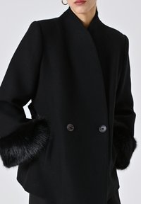 Cappotto double-breasted nero in tessuto strutturato, con bottoni prominenti e morbidi accenti in pelliccia sintetica sui polsini.