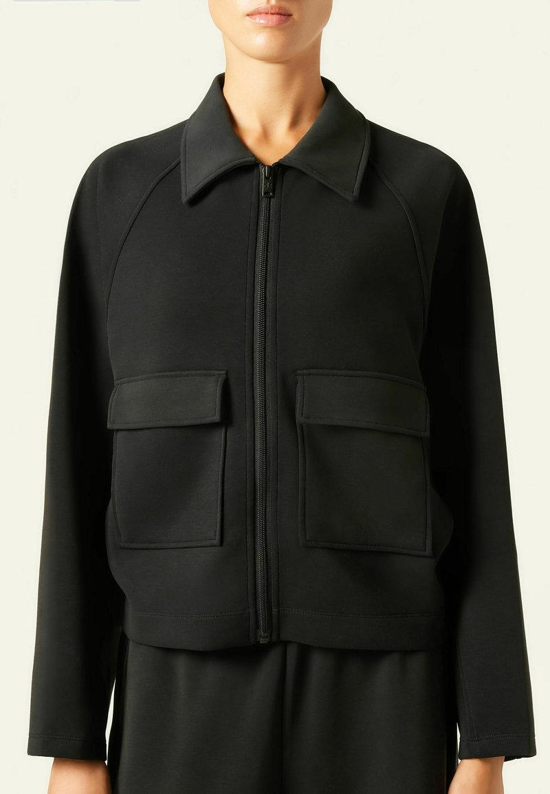 Schwarze taillierte Jacke mit strukturiertem Kragen, Frontreißverschluss und zwei großen aufgesetzten Taschen. Hergestellt aus einem glatten, elastischen Stoff. Normale Passform.