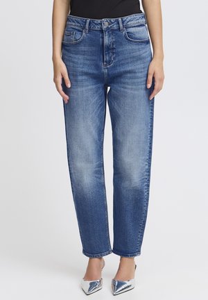 Frau trägt blaue Jeans mit geradem Bein, vorne ausgewaschen, und silberne spitze Absätze, steht vor einem schlichten Hintergrund.