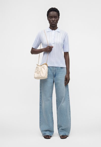 Femme portant un polo bleu clair à manches courtes, un jean bleu clair à jambes larges, des sandales noires, et tenant un sac d'épaule matelassé crème avec une chaîne dorée.