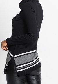 Černý top s dlouhým rukávem a vysokým výstřihem, zdobený šedými a bílými vodorovnými pruhy na lemu a bočních rozparcích. Jemná pletená textura.