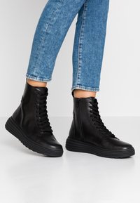 Stivali alti neri in pelle liscia con punta tonda, lacci sul davanti, zip laterale e suola in gomma spessa. Indossati con pantaloni di jeans blu.