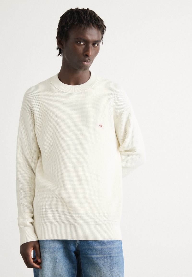 Man in een witte gestructureerde trui met lange mouwen en een klein rood logo op de borst, met blauwe jeans, staande tegen een witte achtergrond.