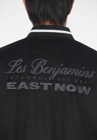 Vista posteriore di una persona che indossa una giacca nera con colletto a strisce bianche e testo ricamato "Les Benjamins Istanbul Est 2011 EAST NOW."