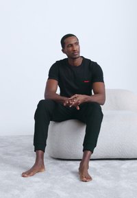 Černé tričko s krátkým rukávem a červeným logem, doplněné černými tepláky. Sedí na světlém, strukturovaném povrchu naboso.