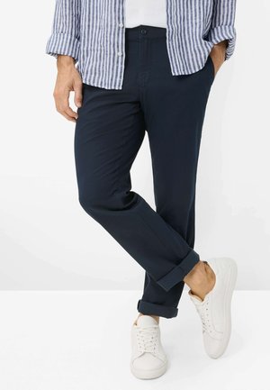 Uomo con pantaloni blu navy, scarpe da ginnastica bianche e camicia a righe blu e bianca con maniche arrotolate, in piedi con le gambe incrociate.