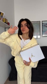 Sárga kapucnis pulóver és hozzá illő, puha anyagból készült nadrág, amint egy fehér dobozt tart, amelyen egy arany Jumpman logó látható, a sötét kanapé h�átterében.