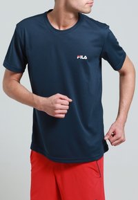 Muž v tmavě modrém sportovním tričku FILA a červených kraťasech pózuje, jako by běžel, na jednoduchém světle šedém pozadí.