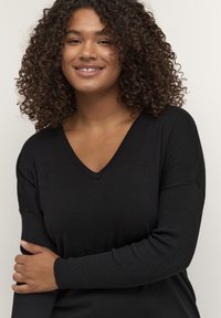 Schwarzer V-Ausschnitt-Pullover mit gerippten Ärmeln, figurbetontem Schnitt und strukturiertem Strick. Die Hand des Models ruht auf ihrem Arm und zeigt den Fall des Stoffes.