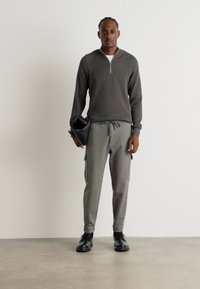 Pulover gri cu dungi, cu fermoar la jumătate și tricou alb pe dedesubt, asortat cu pantaloni cargo verzi și pantofi negri, ținând o geantă neagră.