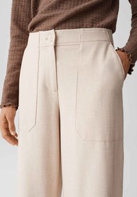 Beige op maat gemaakte broeken met zijzakken, voorzien van een knoopsluiting en een gestructureerde bruine top. De stof lijkt glad en stevig.