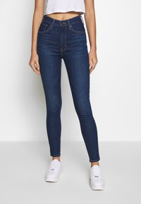 Jeans ajustados de mezclilla azul oscura, con una cintura alta, costuras naranjas y un cierre de botón frontal. Combinados con zapatillas blancas.