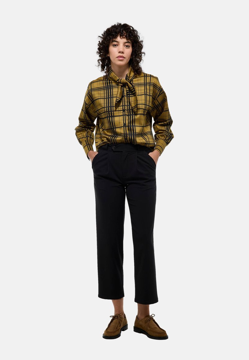 Blouse à carreaux jaunes avec un col nœud papillon, associée à un pantalon noir ajusté et des chaussures marron. Texture lisse et forme ajustée.