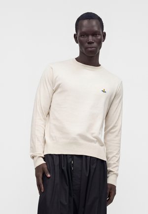 Hombre con suéter beige con un pequeño logo colorido bordado en el pecho y pantalones negros sueltos contra un fondo claro y liso.